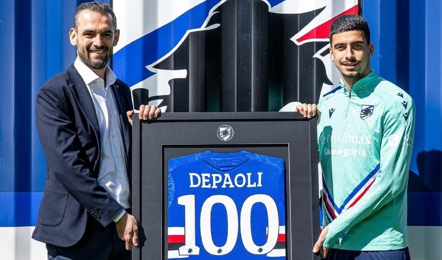 Due uomini, uno vestito elegante e l'altro vestito sportivo, mostrano una maglia speciale della Sampdoria in bacheca col numero 100 sulla schiena. Alle loro spalle un container della Sampdoria.