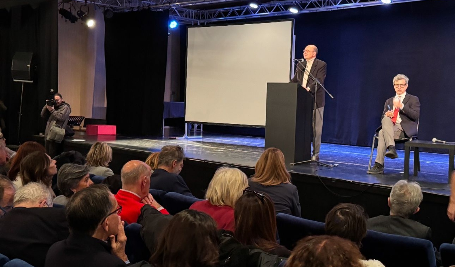 Una sala di un teatro con due persone sul palco, una in piedi e l'altra seduta