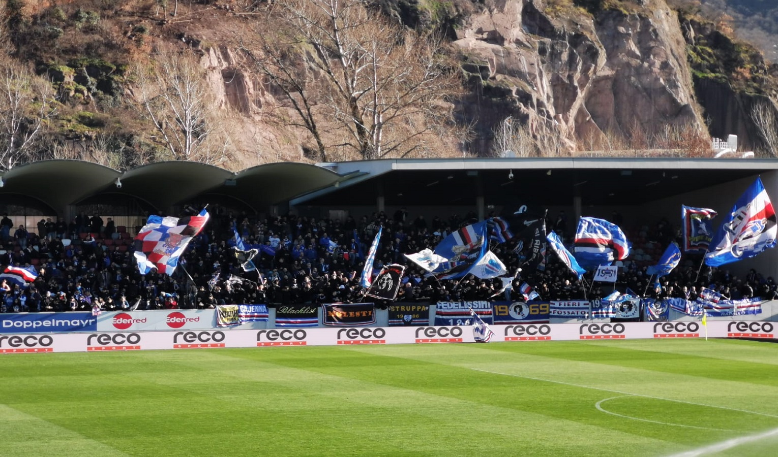 I tifosi della Sampdoria sbandierano in un settore ospiti di uno stadio sotto il sole e sotto un monte